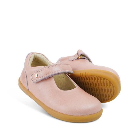 The Bobux Iwalk IW Delight Mary Jane Pink Dusk Pearl features a light pink leather upper, Velcro straps, and tan rubber soles. One shoe stands upright while the other is tilted to display its textured sole.