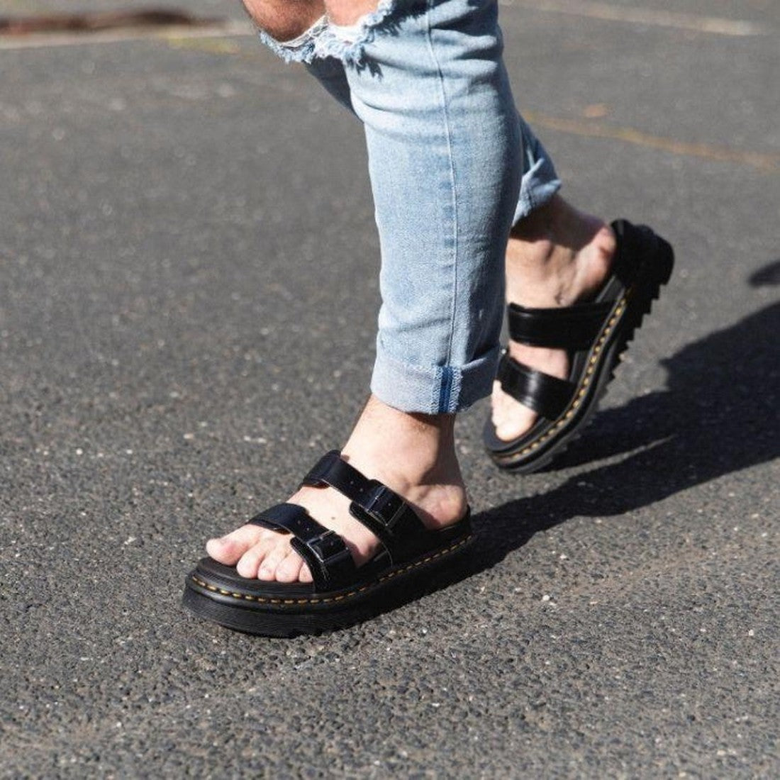 A person walks on a paved surface wearing light blue rolled-up jeans and Dr Martens Black Myles Black Bar Brando Sandals with adjustable buckle straps. Only their lower legs and feet are visible., 190665107562, 190665107579, 190665107586, 190665107593, 19