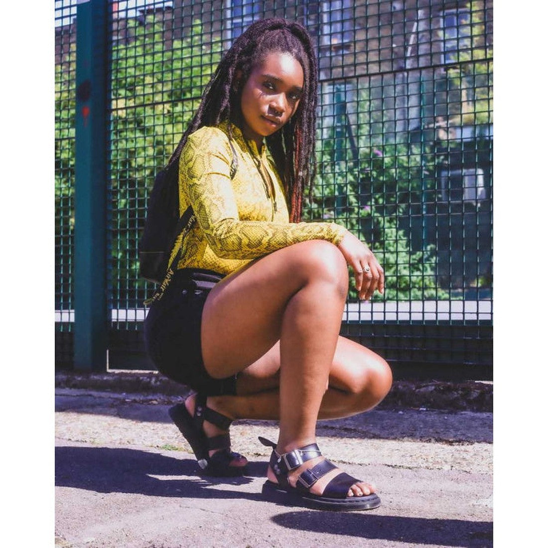A woman with long braids crouches by a fence outdoors, wearing a yellow snake-print top, black shorts, and black Dr. Martens Gryphon Strap Sandal Black Brando, looking confidently at the camera as sunlight creates dynamic shadows., 883985648982, 883985648