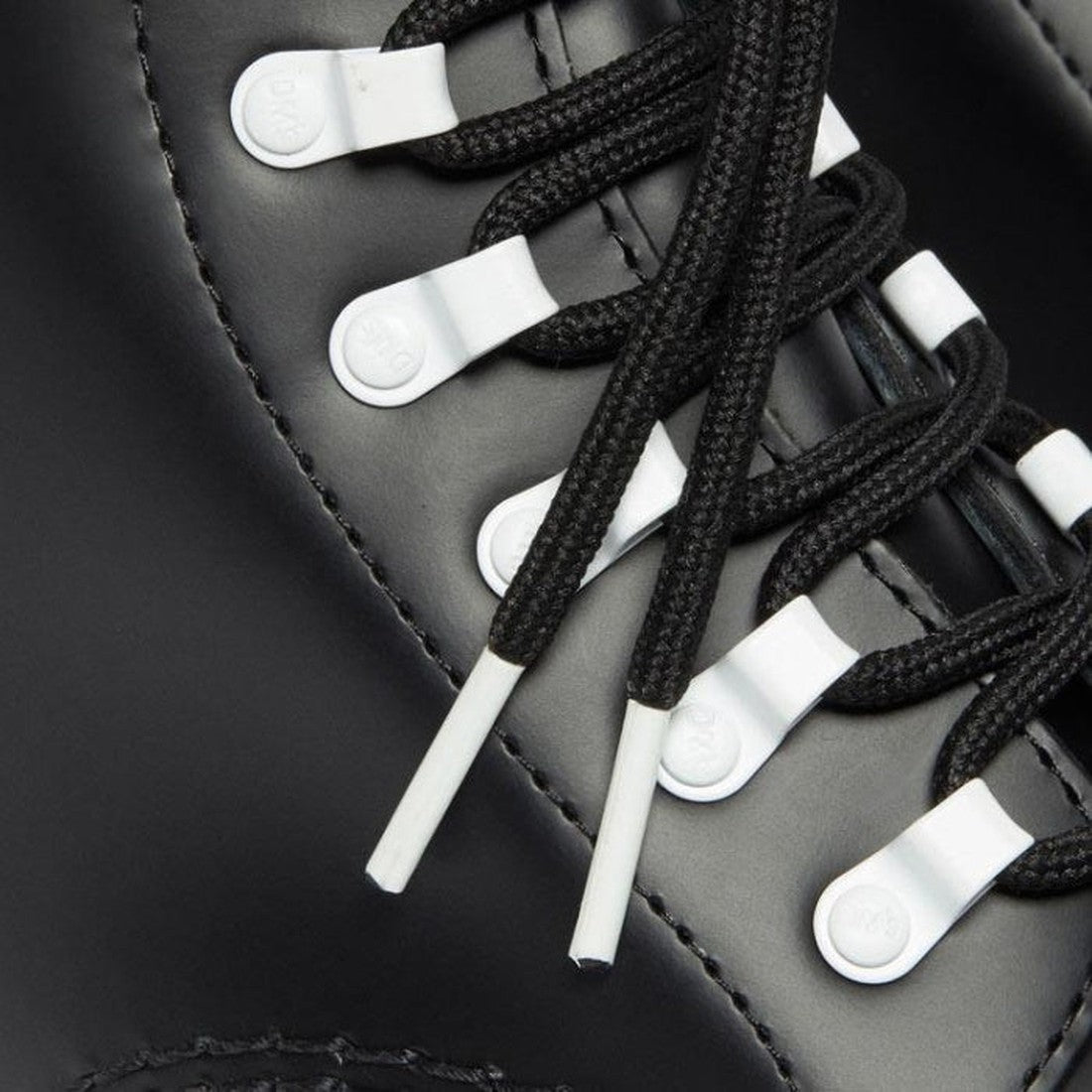 Close-up of black laces threaded through white eyelets on the Dr Martens 1460 Disrupt Hardware 8 Eye Boot Black White, showcasing the lace tips and detailed stitching on its sleek surface reminiscent of smooth leather boots., 22281, 22292, 22293, 19066544