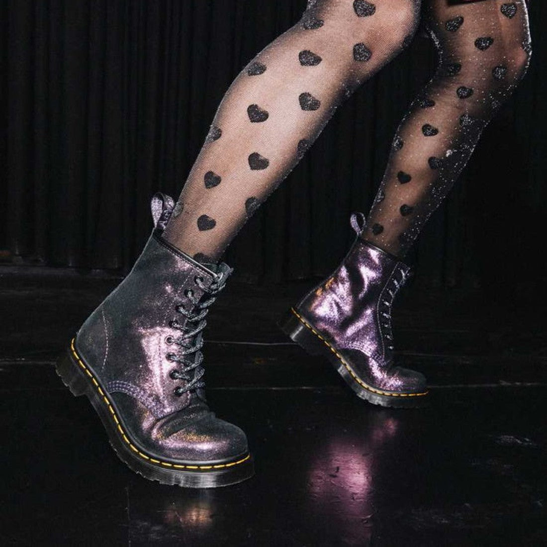 A person wearing sheer black heart-pattern tights and Dr Martens 1460 Disco Iridescent Suede Purple Gold Black boots (wmns 5/uk3) walks on a dark stage with a black curtain backdrop., 190665438895, 190665438901, 190665438918, 190665438925, 22819, 19066543