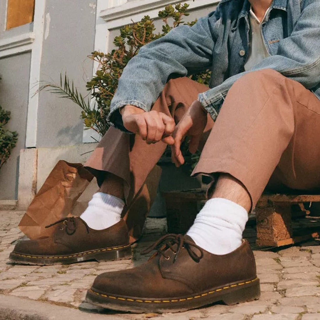 A person in tan pants, a denim jacket, white socks, and Dr Martens 1461 Crazy Horse Oxford Shoe 3 Eye Dark Brown sits on a wooden pallet outdoors with a brown paper bag nearby and greenery in the background., 33438, 800090826708, 800090826722, 80009079813