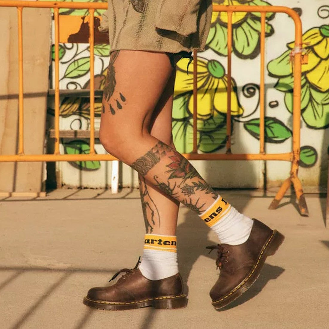 A tattooed person wears Dr Martens 1461 Crazy Horse Oxford Shoe (3 Eye, Dark Brown), white socks with yellow Martens text, and tan shorts while walking past a yellow barrier and a green-and-yellow mural., 33438, 800090826708, 800090826722, 800090798135, 8