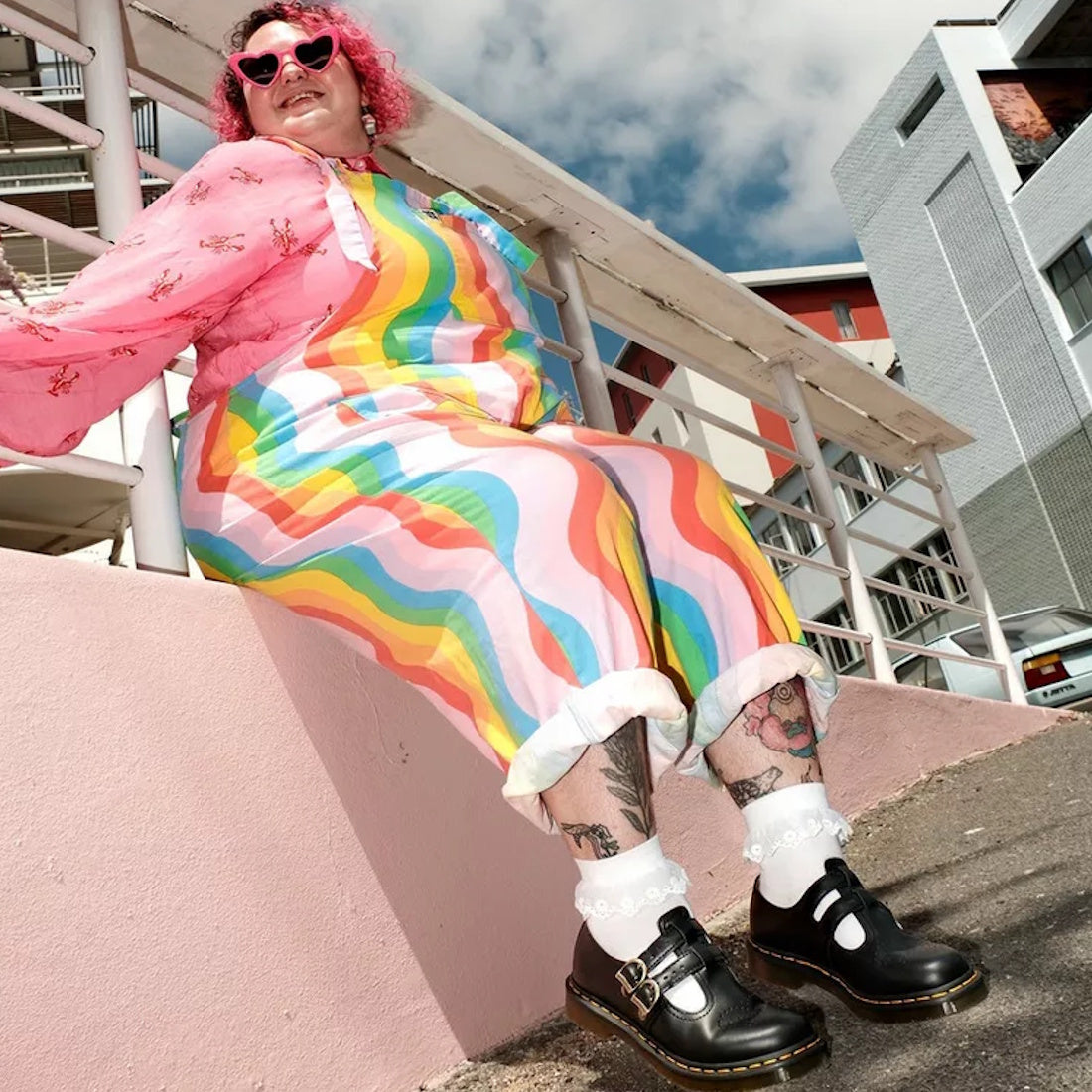 A person with pink hair and heart-shaped sunglasses sits on a pink ledge, wearing a rainbow-striped jumpsuit, lace socks, and Dr Martens 8065 2 Strap Mary Jane Shoe Black Smooth, smiling with modern buildings in the background., 883985051768, 883985051775