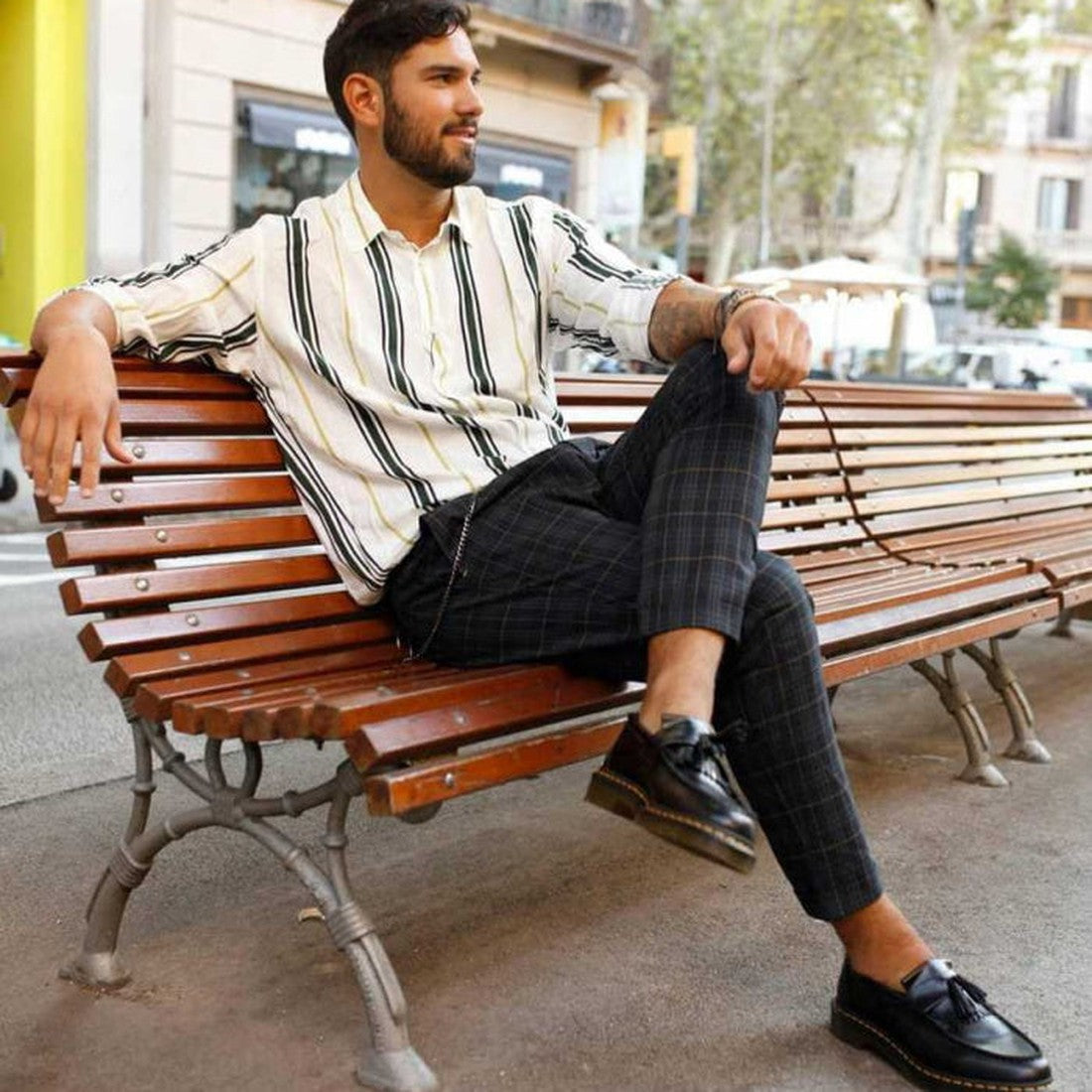 A dark-haired, bearded man relaxes on an outdoor wooden bench, wearing a white striped shirt, plaid pants, and Dr Martens Adrian YS Black Smooth Tassel Loafer Shoes with yellow stitching. Trees and buildings are visible behind him., 22422, 883985989108, 8