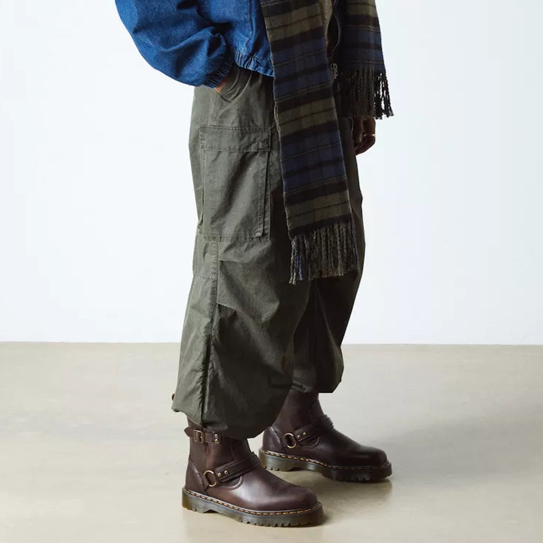 A person wearing Dr Martens Anistone Harness Bex Crazy Dark Brown Horse Biker Boots, olive cargo pants, a blue jacket, and a plaid fringe scarf stands on a light-colored floor against a white wall., 49399, 190665745689, 190665745696, 190665745719, 49407, 