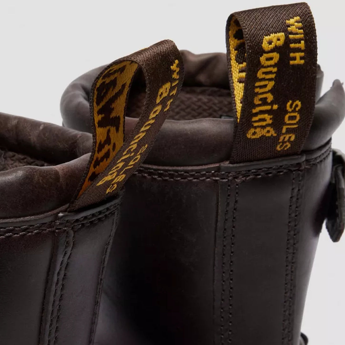 Close-up of the back of two Dr Martens Anistone Harness Bex Crazy Dark Brown Horse Biker Boots, made from Crazy Horse leather, featuring yellow and black pull tabs with "WITH BOUNCING SOLES" in yellow text., 49399, 190665745689, 190665745696, 190665745719