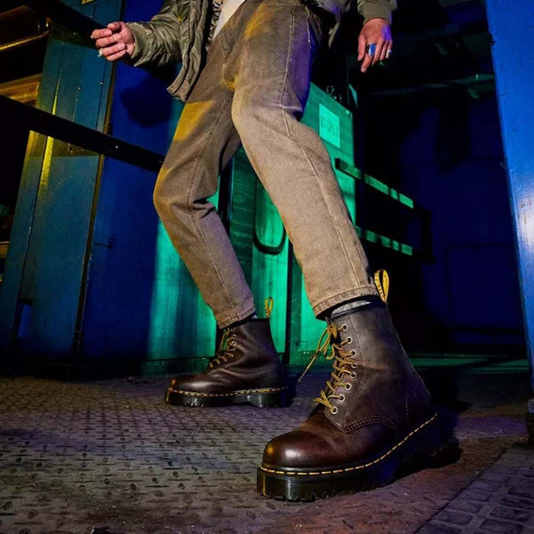 Wearing Dr Martens - Bex 1460 8 Up Leather Crazy Horse Brown boots and tan pants, a person stands on a metal floor in moody green-blue industrial lighting. The low-angle shot highlights the rugged boots against the dramatic setting., 32000, 190665498615, 