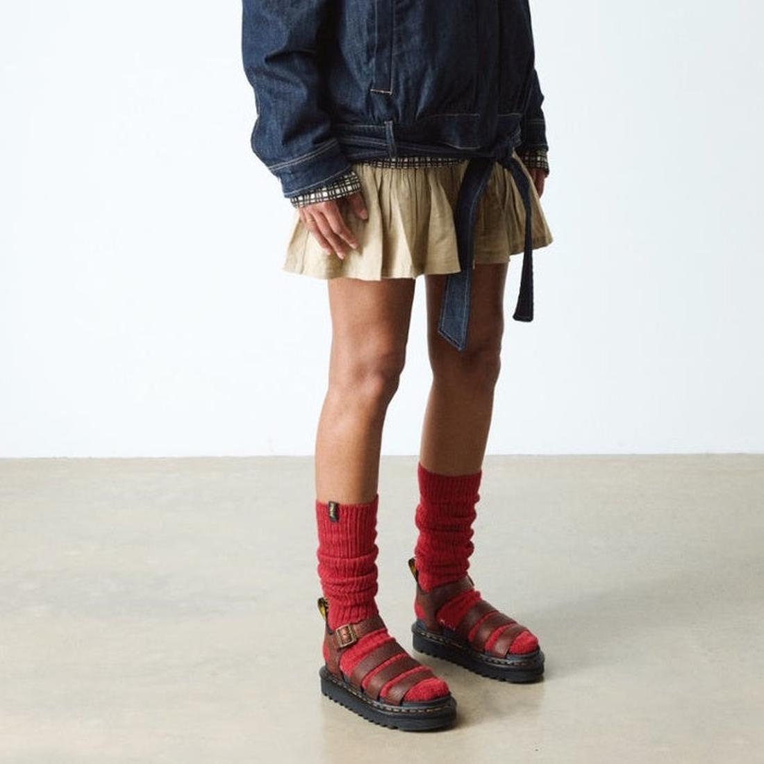 A person in a denim jacket, beige skirt, red slouchy socks, and Dr Martens 3 Strap Blaire Sandal in Cashew Ambassador Leather Tan stands on a smooth light floor against a plain white background., 190665772005, 190665772012, 190665772029, 190665772043, 190