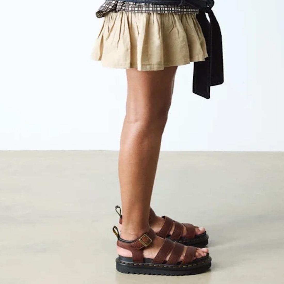 A person wearing a beige pleated skirt and Dr Martens 3 Strap Blaire Sandal in Cashew Ambassador Leather Tan stands on a light floor with a white background, showcasing these stylish, lightweight sandals., 190665772005, 190665772012, 190665772029, 1906657