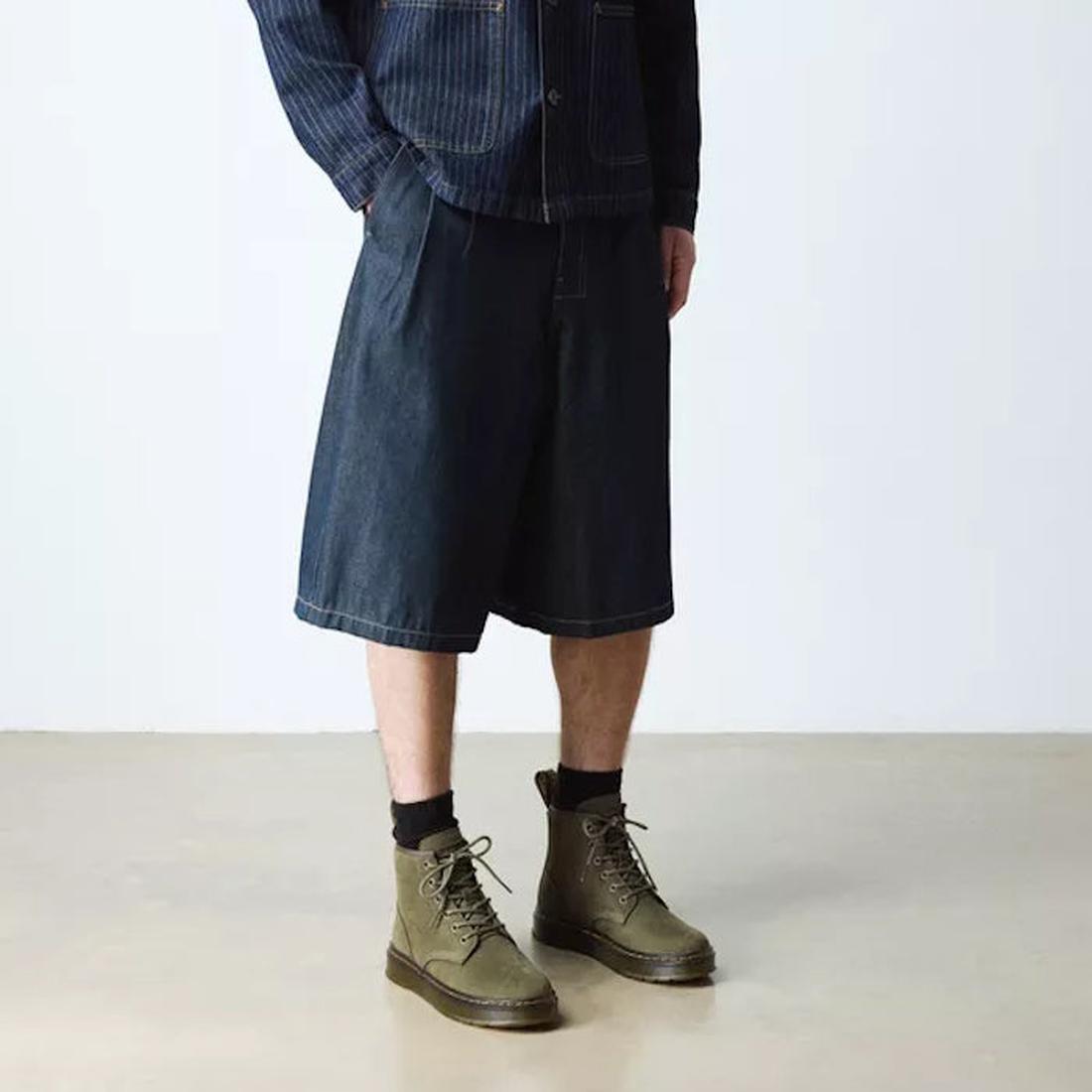 A person wearing a pinstriped jacket, dark knee-length denim shorts, black socks, and Dr. Martens Brookline Chukka Boots in Olive Milled Nubuck Leather with a lightweight outsole stands on a light-colored floor against a plain white background., 51342, 19