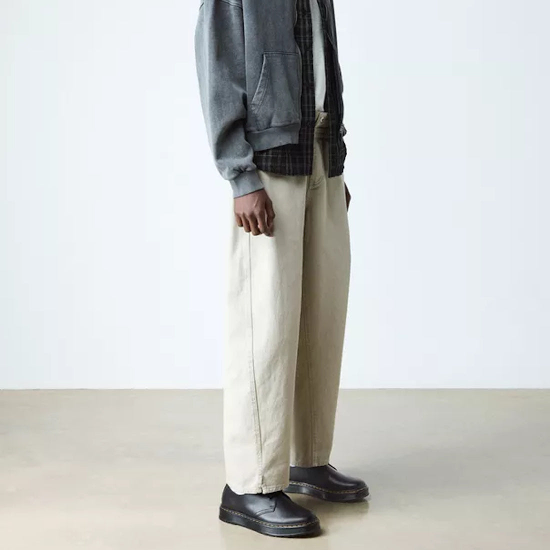 A person wearing a gray jacket, dark shirt, wide beige pants, and Dr Martens Brookline Dark Black Lusso Leather Shoe stands indoors against a plain white background. Only the lower part of their body is visible.