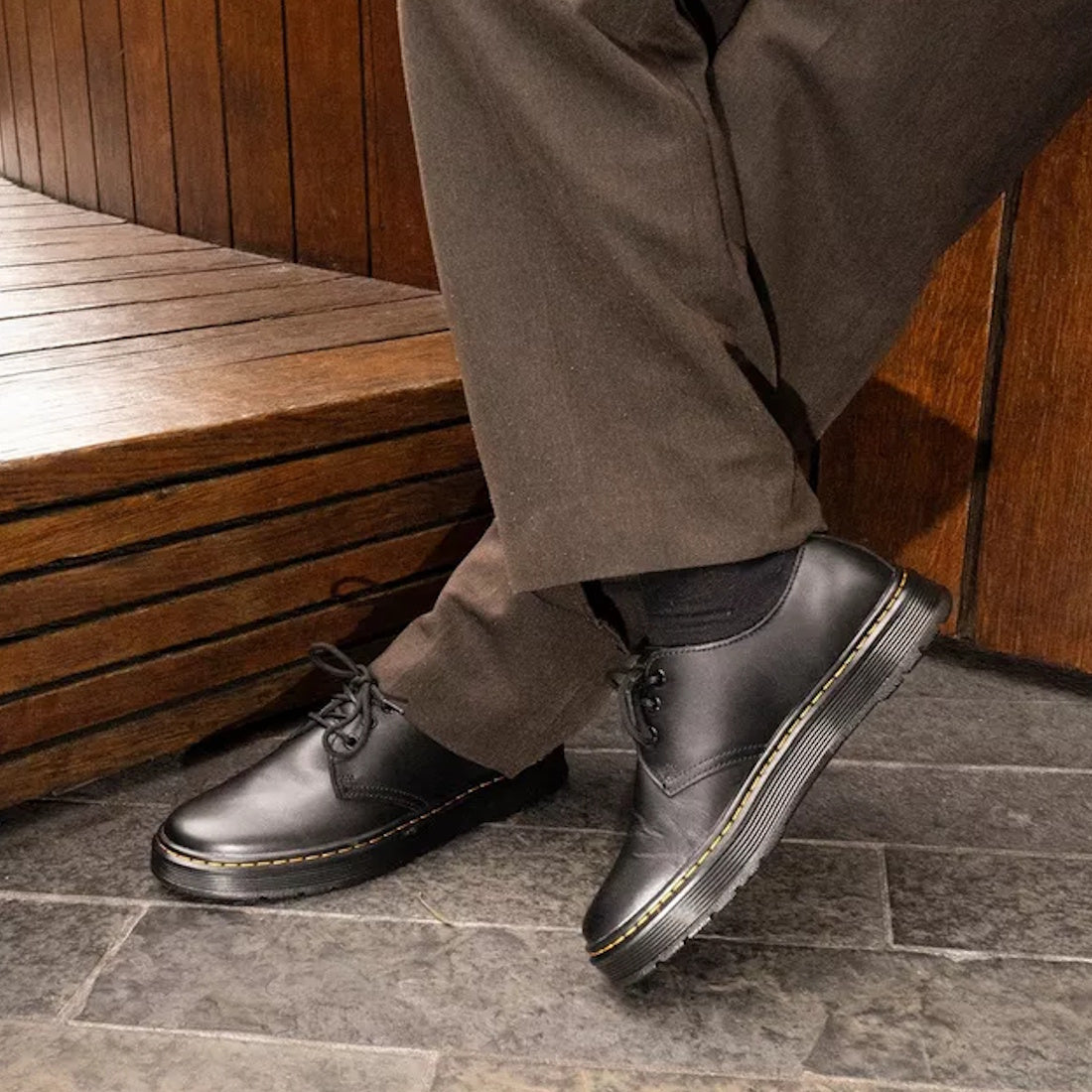 A person in dark brown pants and Dr. Martens Brookline Dark Black Lusso Leather Shoes sits on a wooden bench with legs crossed over gray stone tile flooring.