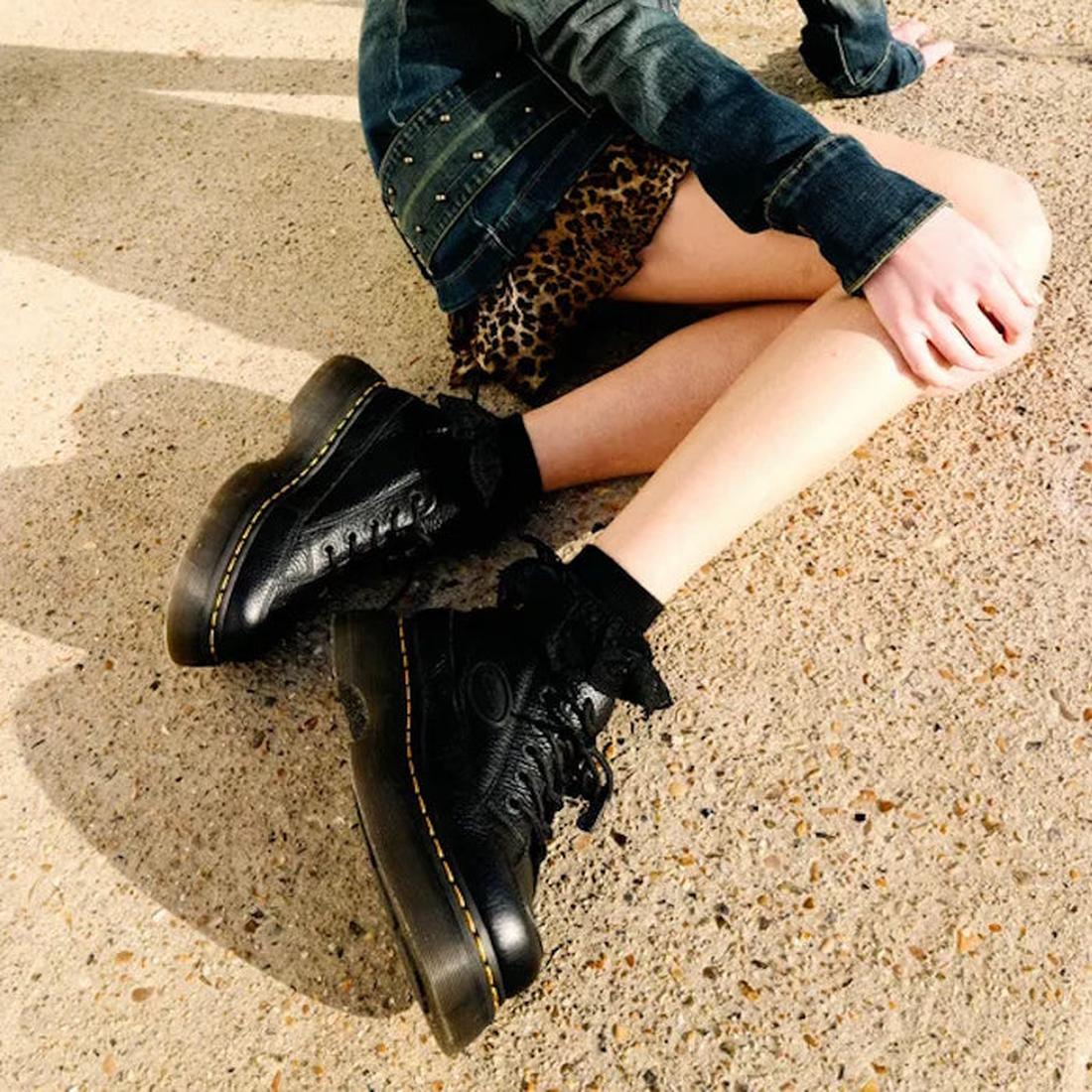Seated on the ground, they wear a denim jacket, leopard print skirt, frilly black socks, and Dr Martens Buzz 5i Milled Nappa Black Leather 5 Eye Shoes, the Goodyear-welted soles catching sunlight on the pavement., 51613, 190665702798, 190665702804, 190665
