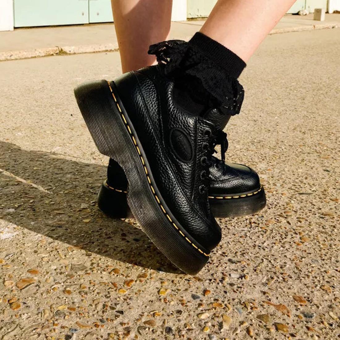 Close-up of a person wearing Dr Martens Buzz 5i Milled Nappa Black Leather 5 Eye Shoes with thick soles, yellow stitching, and black lace-trimmed socks, standing on textured concrete., 51613, 190665702798, 190665702804, 190665702828, 51618, 51619