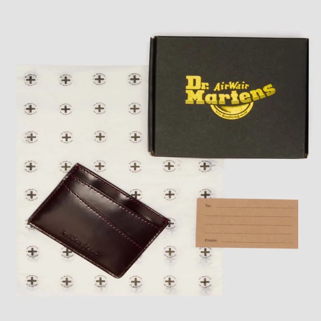 A Dr. Martens gift set features the Card Holder II in Cherry Red Oxford Brush Vegan, a branded black box, anchor-patterned wrapping paper, and a tan gift tag with To and From fields.
