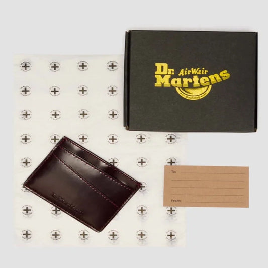 A Dr. Martens gift set features the Card Holder II in Cherry Red Oxford Brush Vegan, a branded black box, anchor-patterned wrapping paper, and a tan gift tag with To and From fields.