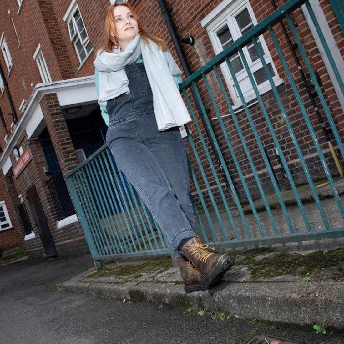 A cheerful woman in overalls relaxes on a blue fence, sporting Dr Martens Doc Crewson Classic Brown Boots with sturdy soles and easy lace-up closure.  ksnz, 190665608861, 190665608878, 190665608885, 190665608908, 190665608915, 190665608922, 190665608946