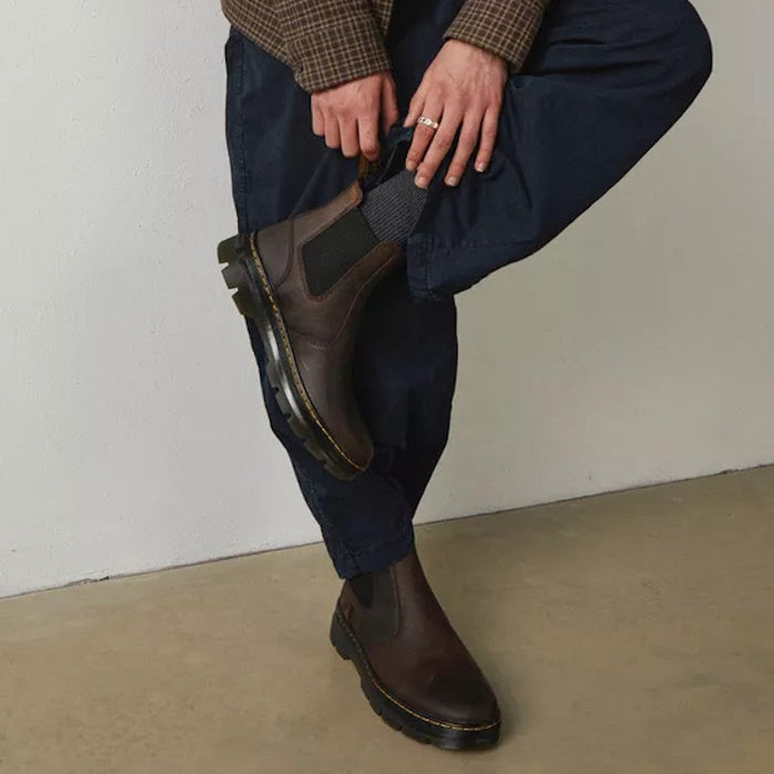 A person leaning against a wall with one leg angled against the others knees, wearing Dr Martens Embury boots in crazy horse brown showing the black elastic gussets,  and yellow stitching around the sole .,
190665358643,
190665358650,
190665358674,
190665