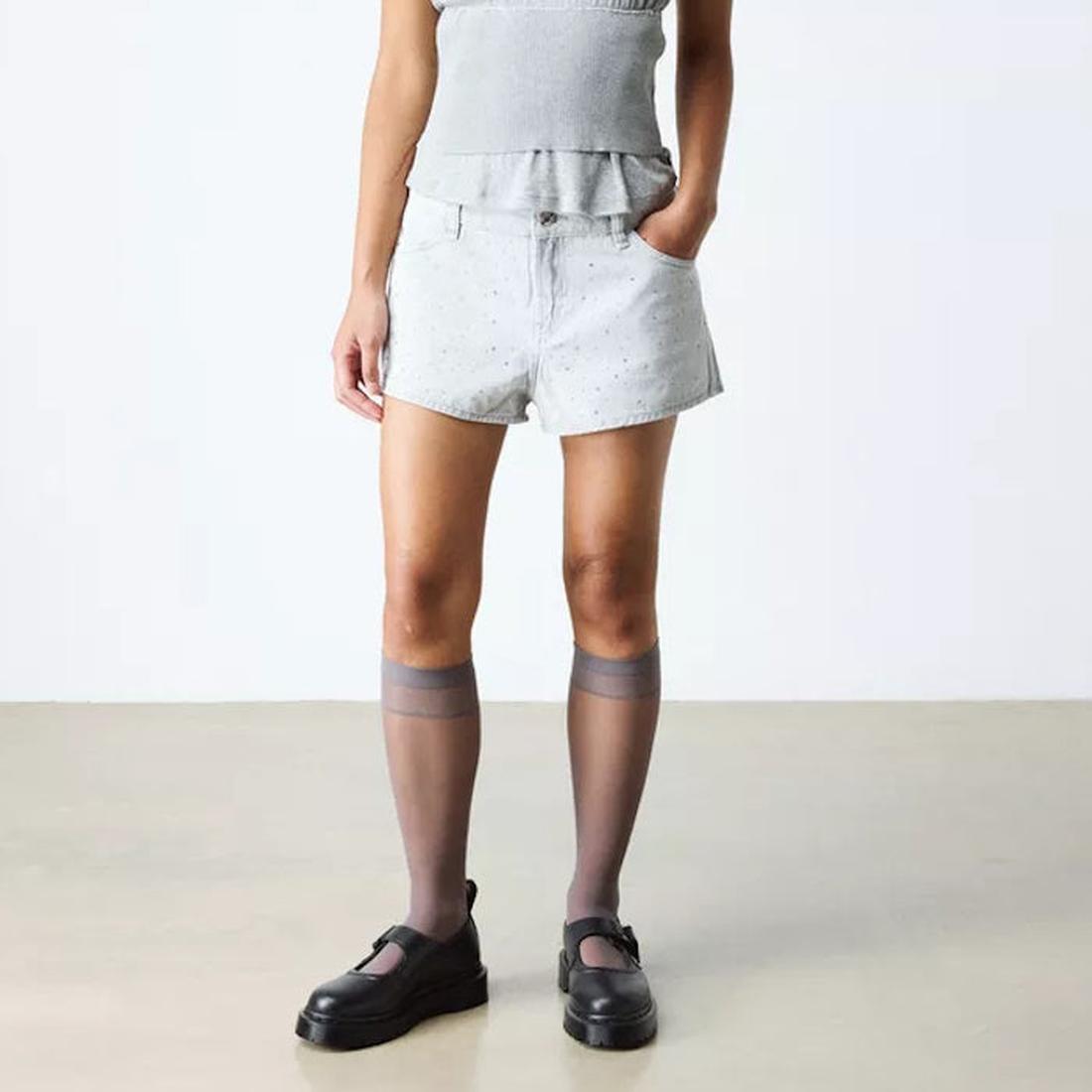 A person wears light gray shorts, a matching top, sheer knee-high socks, and Dr Martens Bex Indica II Mary Jane Tech Nappa Mono Black shoes against a plain background. With one hand in their pocket, their face remains out of view., 190665804744, 190665804