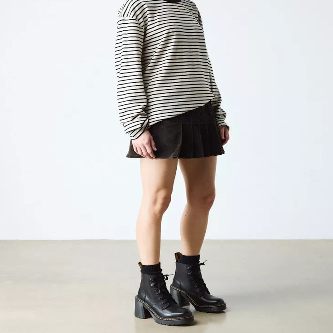 A person in a black-and-white striped long-sleeve shirt, black pleated skirt, black socks, and Dr Martens Jesy Sendal 6 Tie Flared Heel Boots stands against a plain background, with their face not visible., 190665511796, 190665511802, 190665511819, 190665