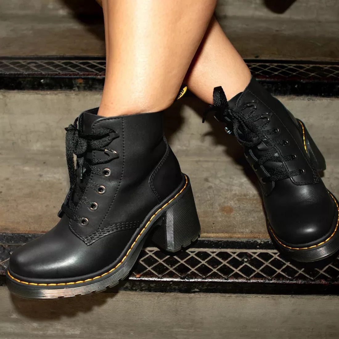 A person wearing Dr Martens Jesy Sendal 6 Tie Flared Heel Boots with chunky heels, yellow stitching, and a Goodyear welt stands on metal and concrete stairs., 190665511796, 190665511802, 190665511819, 190665511833, 190665511840, 190665511857