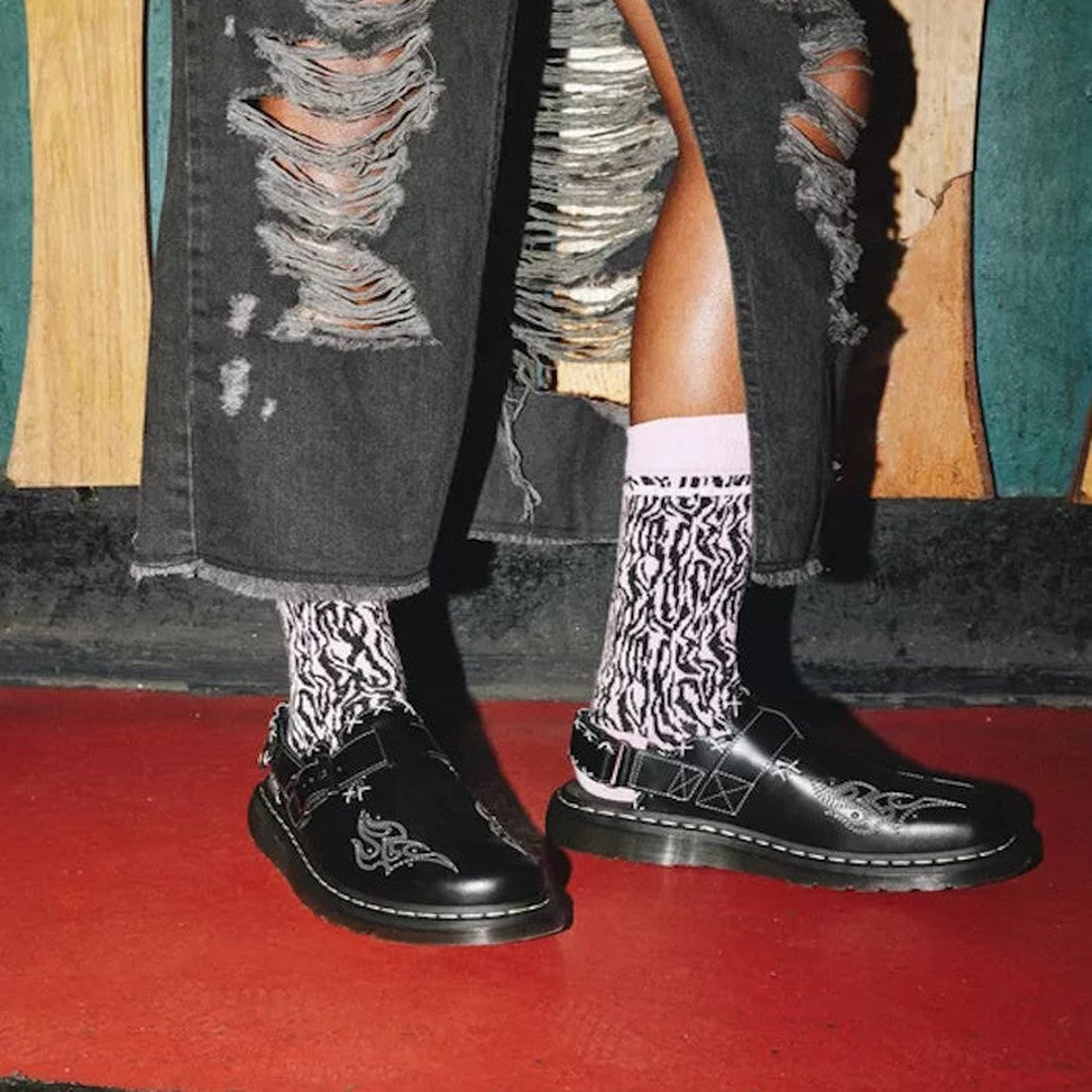 A person wearing Dr Martens Black Jorge II Gothic Americana Leather Slingback Mules, pink patterned socks, and black ripped jeans stands on a red floor in front of a colorful wooden wall., 35783, 190665643480, 190665643497, 190665643503, 35797, 1906656435