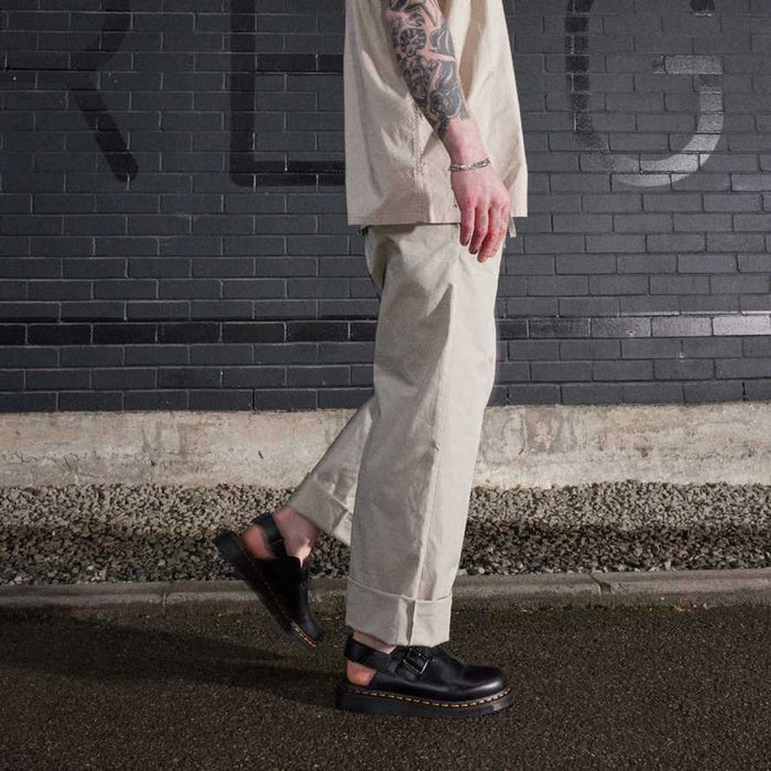 A person wearing cuffed beige pants, a beige t-shirt, and Dr Martens Black Jorge II Brando Sling Back Sandal Covered Toe Mules stands on a wet street in front of a black brick wall. Their arm displays visible tattoos., 29216, 190665560824, 190665560831, 1