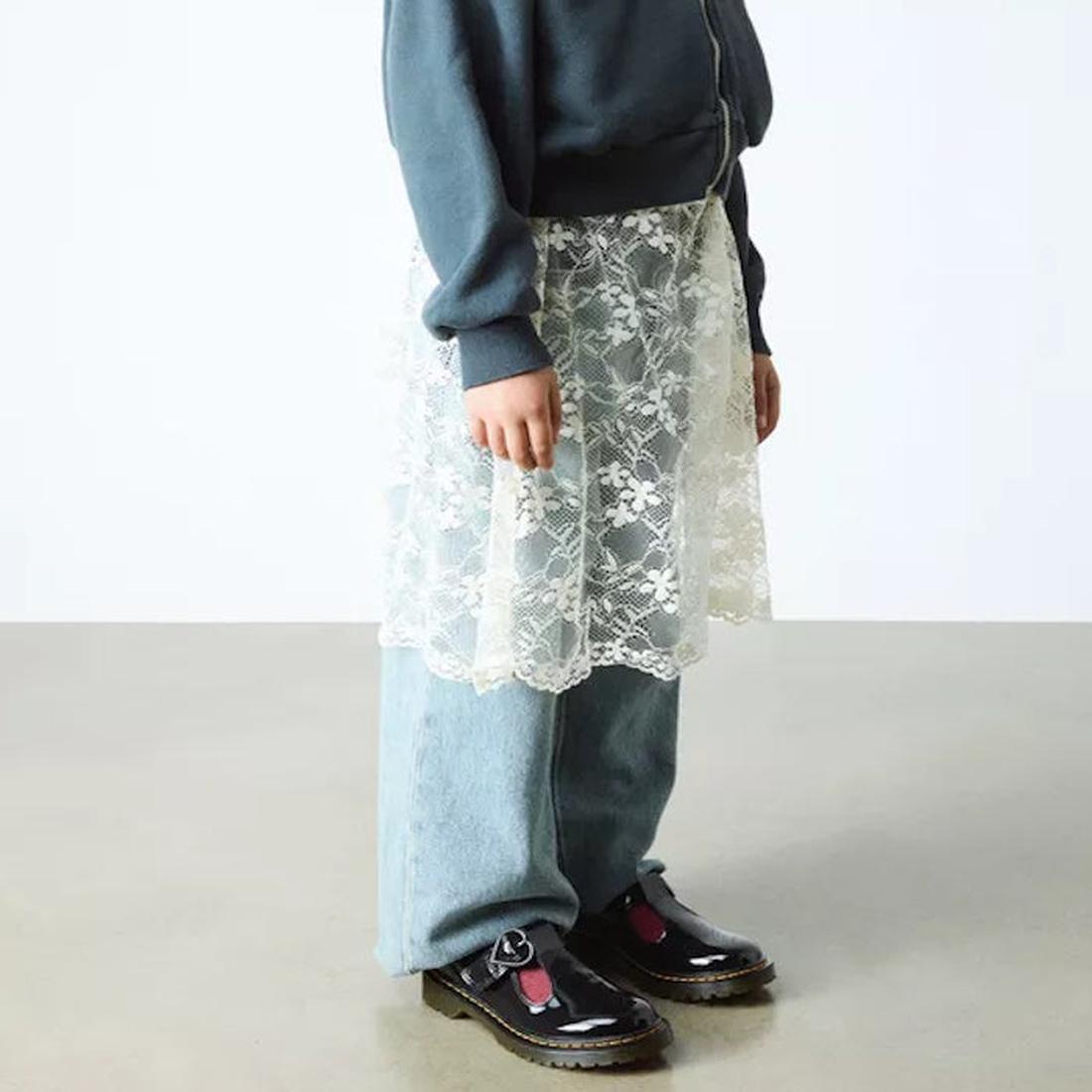 A person stands indoors on a polished floor wearing light blue jeans, a dark blue jacket, a sheer white lace skirt overlay, and Dr Martens kids - Polley II Heart T Bar Junior Black Patent shoes. The photo is cropped at the shoulders., 50432, 50433, 190665