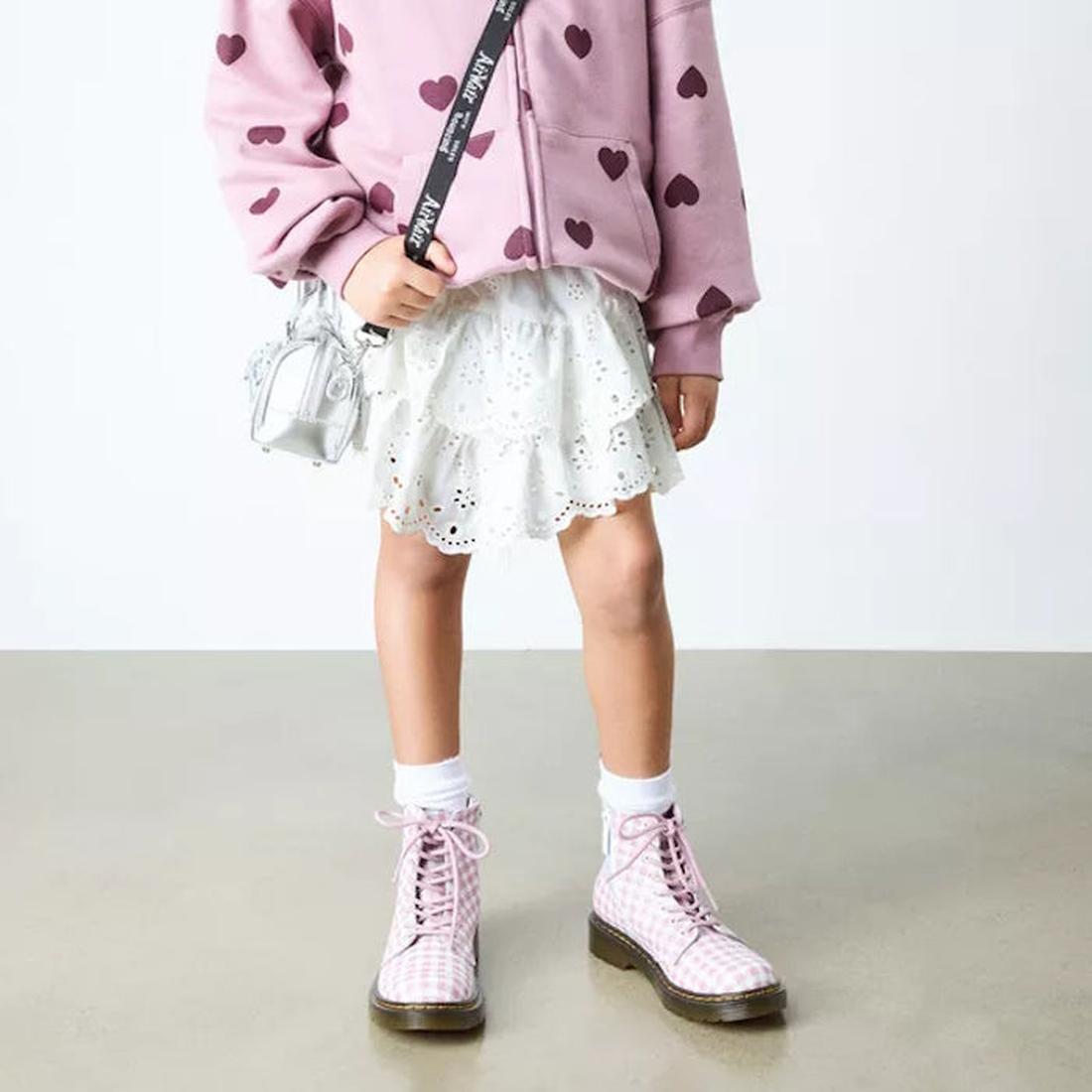 A child in a pink heart-print sweatshirt, white eyelet skirt, white socks, and Dr Martens kids - 1460 J Juniors Gingham Pink Lace Up Boots with zip stands on a light floor with an Athena leather crossbody bag against a plain background., 50424, 50425, 504
