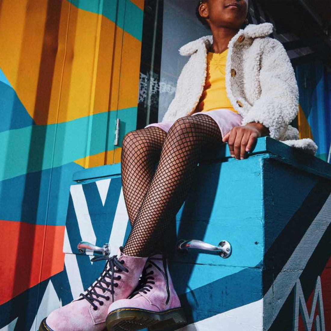 A child in a white fuzzy jacket, yellow dress, fishnet tights, and Dr Martens Kids 1460 J Junior Tie Dye Pink Suede Boots sits on a blue box before a vibrant geometric mural., 190665493597, 190665493610, 190665493634, 27417, 190665493542, 190665493566, 19