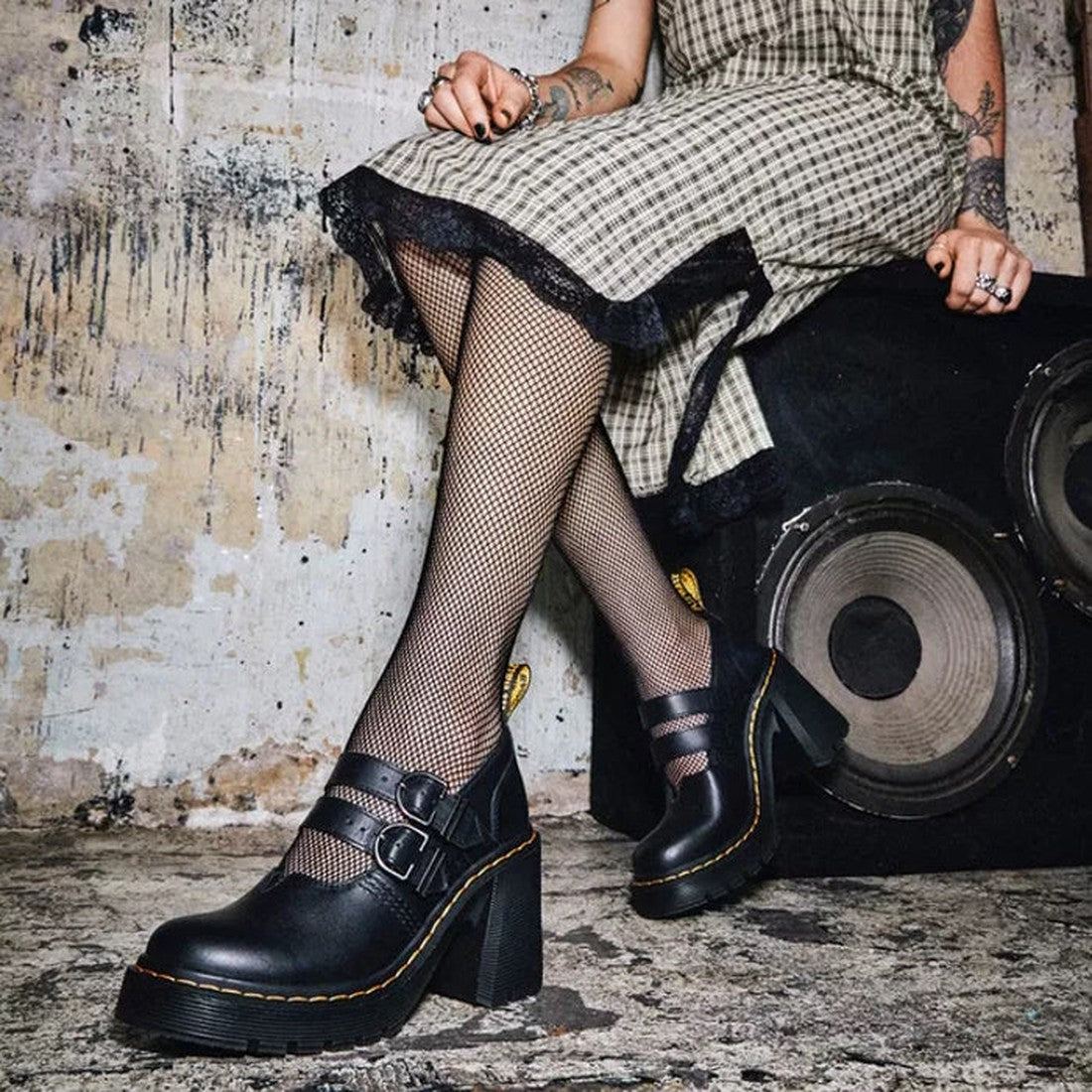 A person in a plaid dress, fishnet tights, and Dr Martens Women's Lottee Eviee Mary Jane shoes with double strap buckle sits on a speaker against a worn wall, showing tattooed hands decorated with rings., 190665459623, 190665459630, 190665459647, 19066545