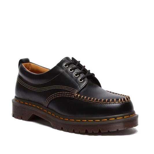 The Dr Martens Lowell Moc Toe Black Leather Shoe features thick brown stitching, a chunky sole, Goodyear welt construction, and yellow edge detailing, shown from an angle highlighting the side and front.