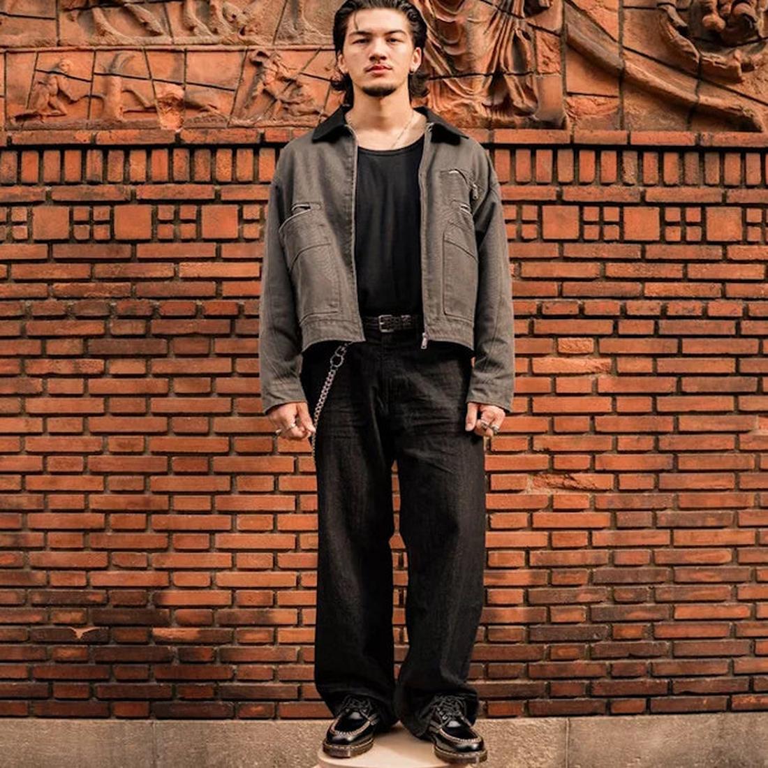 A man stands before a decorative brick wall, wearing a black t-shirt, gray jacket, loose black pants, and Dr Martens Lowell Moc Toe Black Leather Shoes with a chain detail. His hands are at his sides as he looks straight at the camera., 50395, 50396, 5039