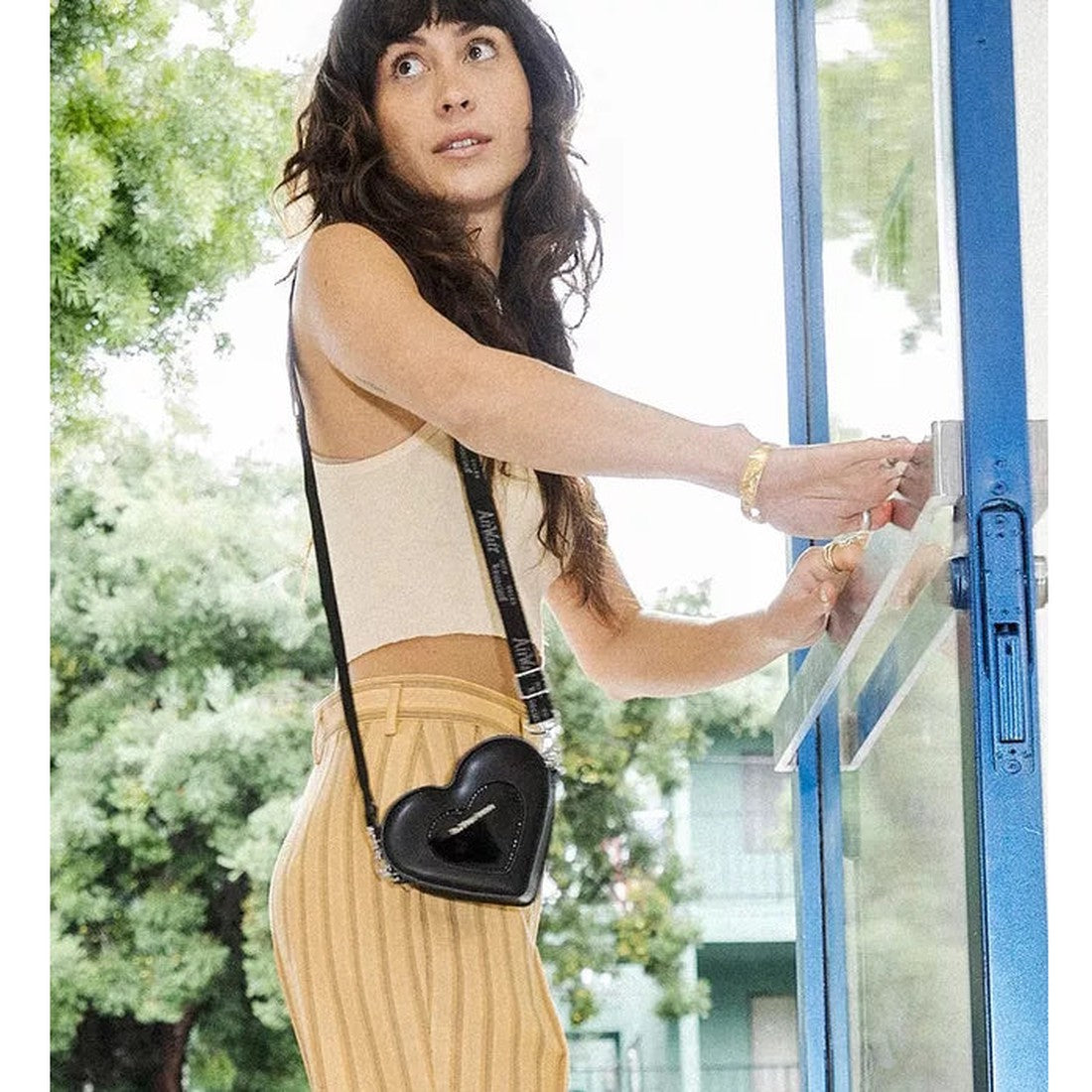 A woman with long dark hair, in a beige crop top and yellow striped pants, opens a blue glass door while carrying the Dr Martens Mini Heart Leather Cross Body Black Patent Leather Satchel. Green trees and a building are visible outside., 190665640373