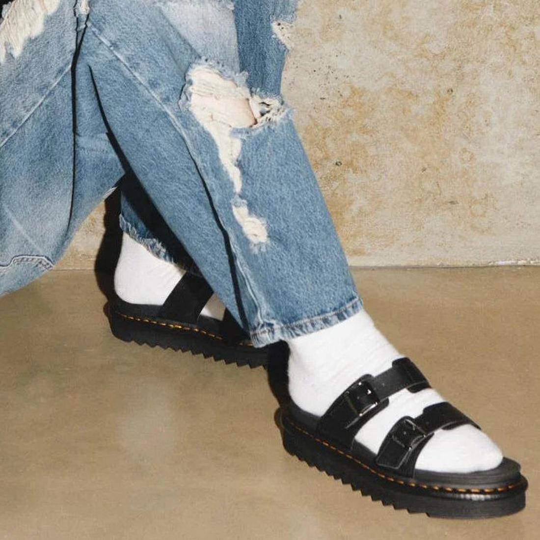 A person sitting on the ground, only the legs and feet in the shot, wearing blue denim jeans , white socks and Dr Martens Myles black leather slide sandals with 2 straps across the foot with buckle on the outer foot for adjustment.,
190665653779,
19066565