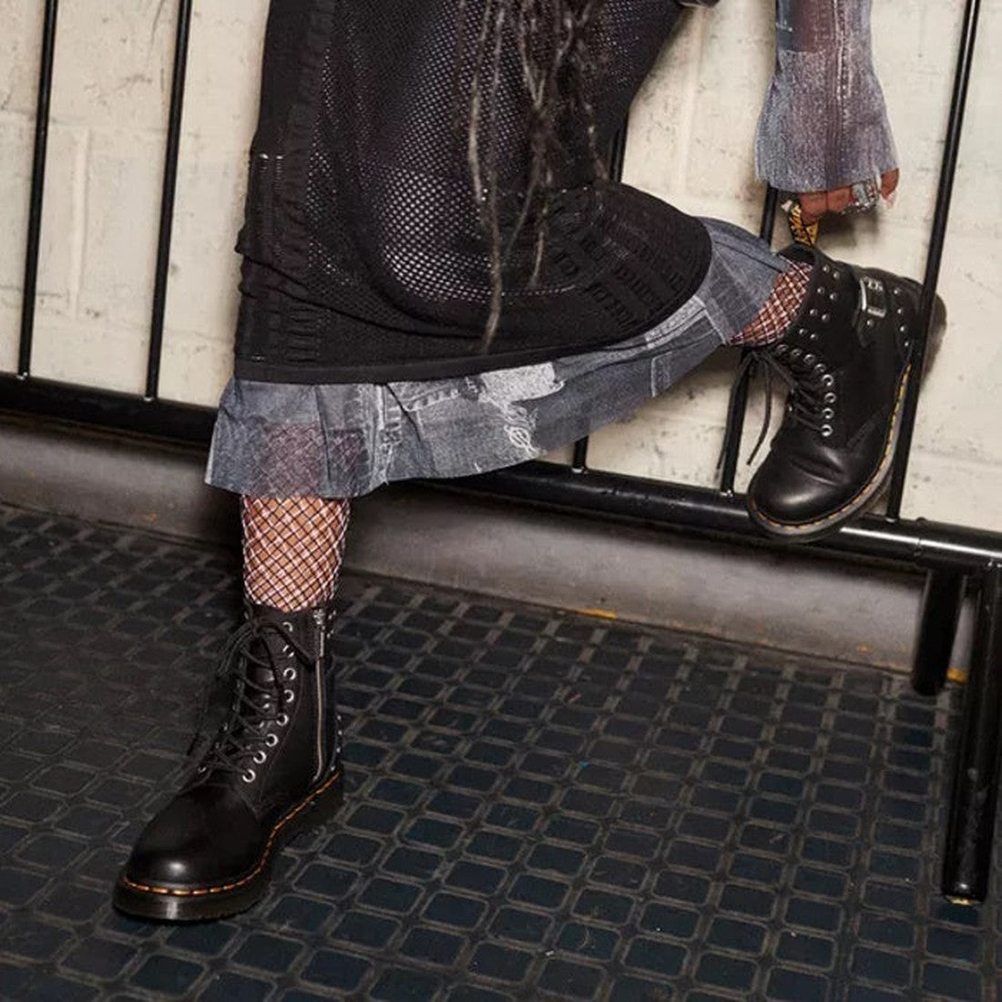 Wearing black Dr Martens 1460 HDW Pascal Hardware 8 eye Nappa boots, fishnet socks, and a layered skirt, a person steps forward on a dark textured floor by a metal railing., 190665552911, 30444, 30445, 190665552928, 190665552935, 190665552942, 19066555295