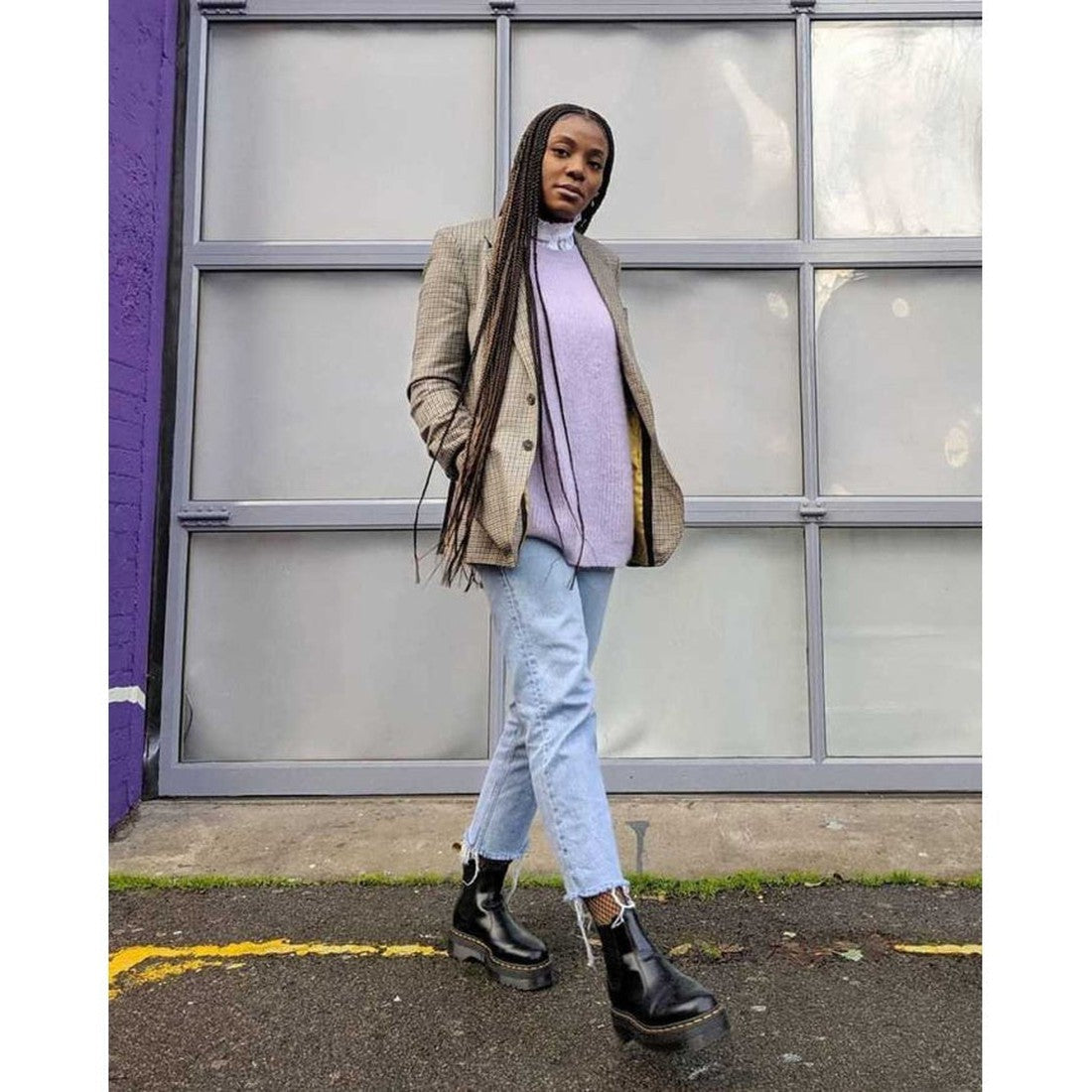 A woman in a plaid blazer, lavender turtleneck, light blue jeans, and Dr Martens - 2976 Quad Chelsea Black Boot stands confidently on pavement with one hand in her pocket, posed against a modern glass-paneled wall., 190665239621, 190665239638, 19066523964