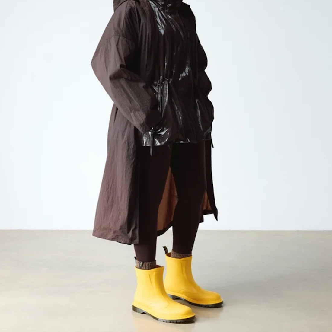 A person wears a long dark brown raincoat with a shiny upper, dark pants, and bright yellow Dr Martens 1460 Sub Max Pull On Rain Boot Yellow PVC, standing indoors against a plain light background., 49435, 190665746464, 190665746471, 190665746495, 49440, 4