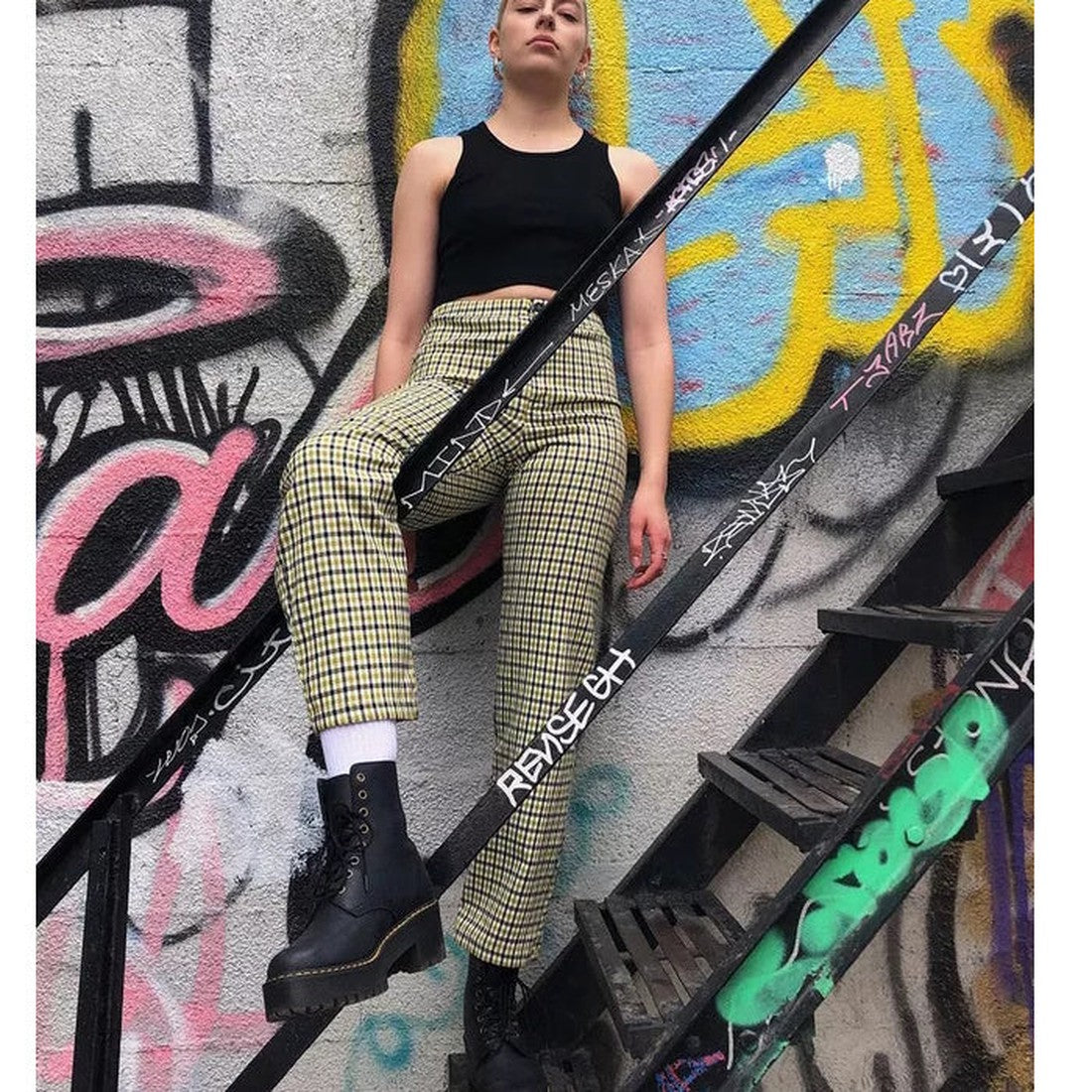 A person in a black tank top, yellow plaid pants, and Dr Martens Women's Shriver Hi Lace Up Leather Boot Black stands on graffiti-covered metal stairs, one foot up, with a colorful graffiti wall behind., 190665158588, 190665158595, 190665158601, 190665158