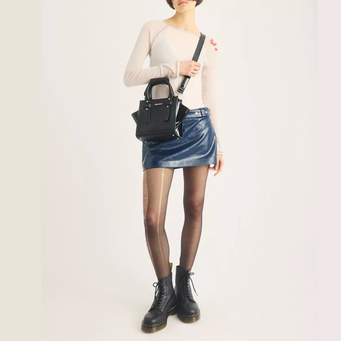 A person wearing a fitted white top, blue mini skirt, sheer tights, and black lace-up boots crafted from sleek black Kiev leather stands against a plain light background, carrying the Dr Martens Small Keiv Leather Tote Bag Black over their shoulder.