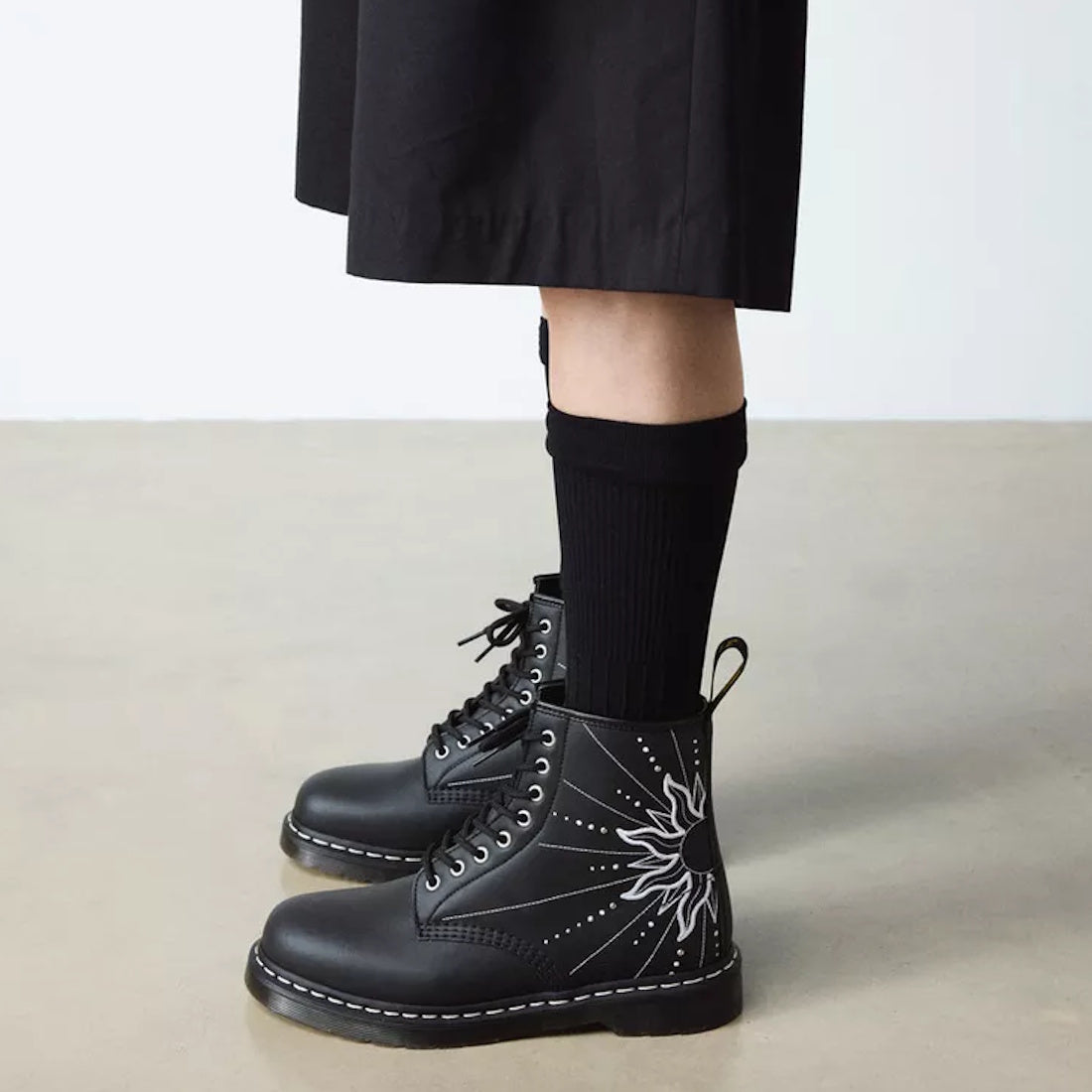 A person in a black skirt and socks wears Dr Martens - 1460 Genix Solar Flare Black 8 Up Eye Boots, featuring a white sunburst design, standing on a light-colored floor against a plain background., 190665742824, 190665742831, 190665742848, 190665742862, 1