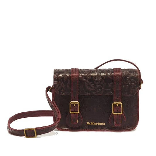A cherry red Dr. Martens 7 Inch Satchel, crafted from Crazy Horse heavyweight leather with embossed floral detailing, featuring a long strap, gold buckles, and “Dr. Martens” lettered in gold on the front.