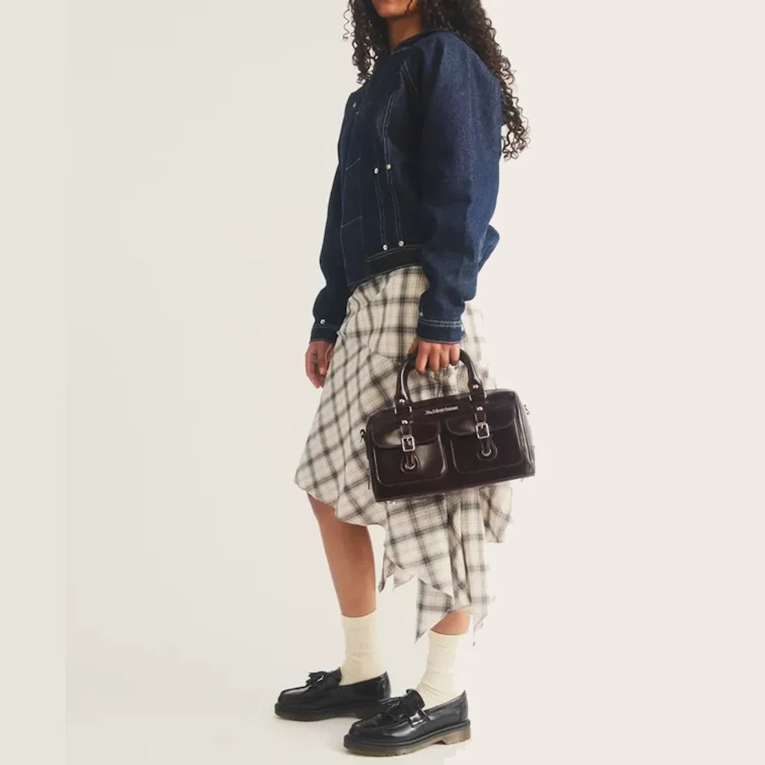A person wearing a dark denim jacket, a plaid asymmetrical skirt, white socks, and black loafers holds the Dr Martens Top Handle Bag in Cherry Red Oxford Rub Off. The background is plain and light.