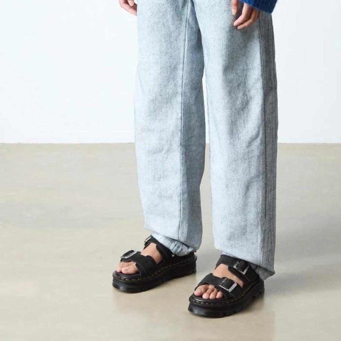 A person stands on a light floor, wearing loose light-wash jeans and Dr Martens Zebzag Black EH Suede 2 Strap Sandals with Back Strap. Only their lower legs and feet are visible., 190665763737, 190665763744, 190665763751, 190665763775, 190665763782, 19066
