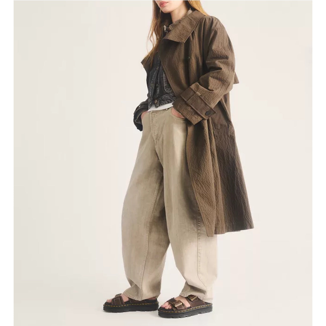 A person with long hair wears a brown textured trench coat, wide beige pants, a patterned shirt, and Dr Martens Zebzag Crazy Horse Brown 2 Strap Sandals with an air-cushioned platform sole, standing against a plain light background., 52549, 190665818604, 