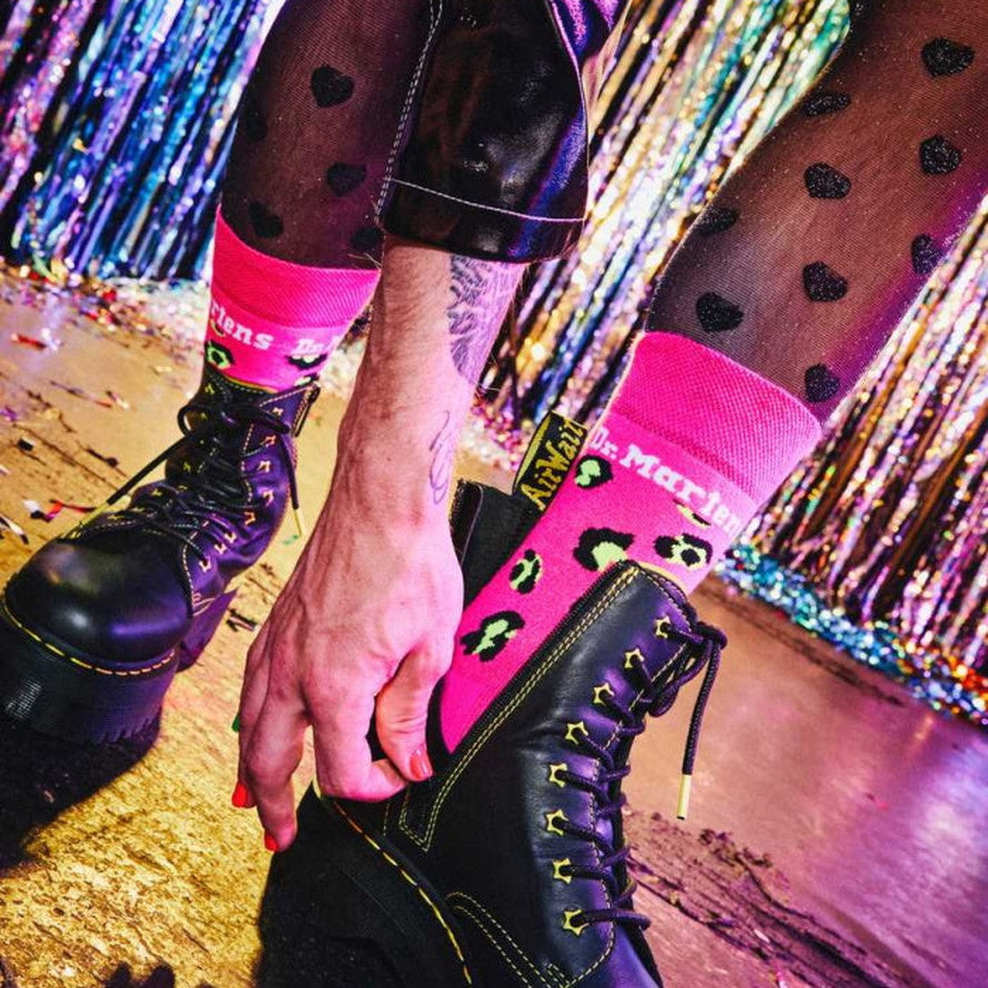 A young person in heart-print pants tries on Doc Martens Black Lace-Up Boots with sturdy soles and pink socks, near festive streamers.  ksnz, 190665515435, 190665515428
