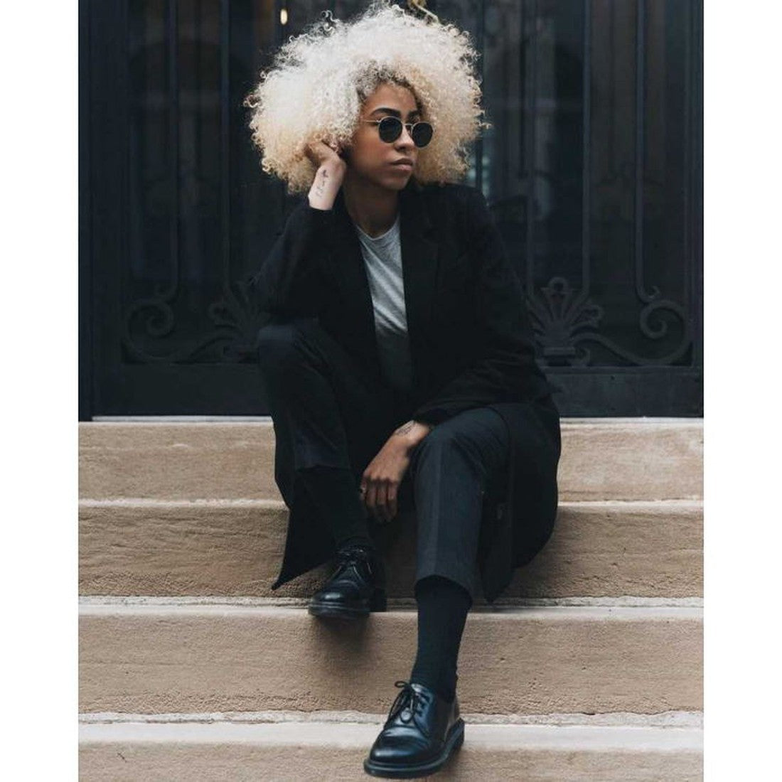 A person with bleach-blond curly hair, sunglasses, a black coat, and black pants sits on outdoor steps wearing Dr Martens 1461 Mono Black Smooth Shoe Oxford Lace Ups, resting one hand on their head and looking to the side., 883985393523, 883985393547, 883