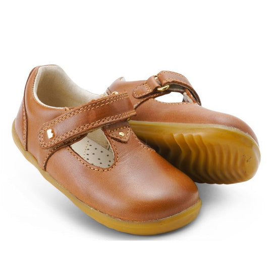 The Bobux Step Up T-Bar Louise Tan Caramel 728318 are toddler shoes in brown leather with Velcro straps and rubber textured soles, shown with one upright and the other on its side to display the sole.