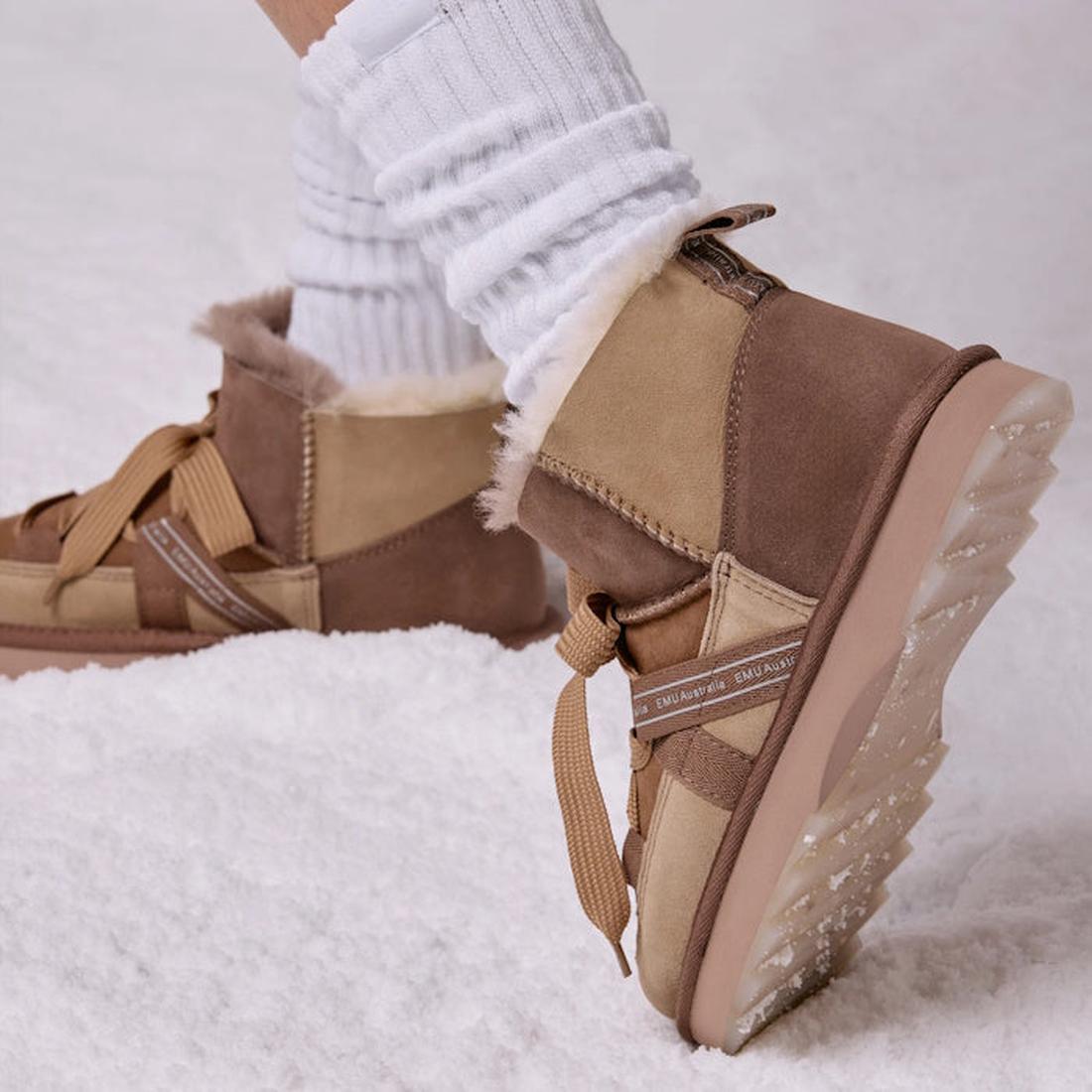 Close-up of a person wearing EMU Women's Blurred Micro Patchwork Sheepskin Boot in Mushroom, featuring tan and brown Australian sheepskin, visible fur lining, thick soles, and ribbon-like laces with white scrunched socks on snow., 47429, 9330071978207, 93